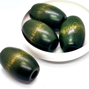 May include: Four oval-shaped beads with a dark green finish and visible wood grain texture. Three beads are clustered inside a small white bowl, while one bead rests outside. Each bead features a central hole for threading, showing a smooth surface.