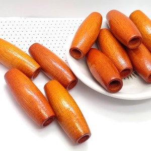 Peut inclure: Un groupe de dix perles en bois de couleur orange chaud. Les perles sont cylindriques et ont une finition lisse et polie. Elles sont disposées sur une assiette blanche.
