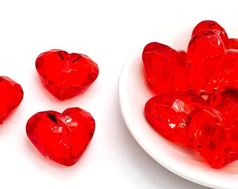 May include: A collection of red heart-shaped beads. Some beads are in a white bowl, while others are scattered on a white surface.