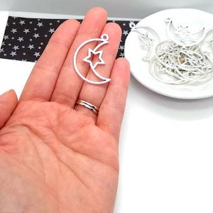 May include: Silver metal charms in the shape of a crescent moon with a star inside. The charms are on a white plate and a hand is holding one of the charms.