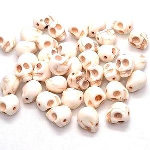 May include: A collection of small, off-white skull-shaped beads featuring brown veining patterns. Each bead has carved eye sockets and a nasal cavity, with a visible hole drilled through the side for threading, scattered across a plain surface.