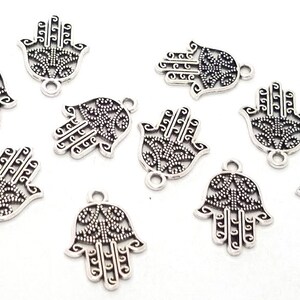 Puede incluir: Un conjunto de nueve dijes de metal plateado con forma de mano de hamsa. Cada dije presenta un dise&ntilde;o intrincado de filigrana calada con un patr&oacute;n decorativo central y una peque&ntilde;a anilla circular en la parte superior para su fijaci&oacute;n.