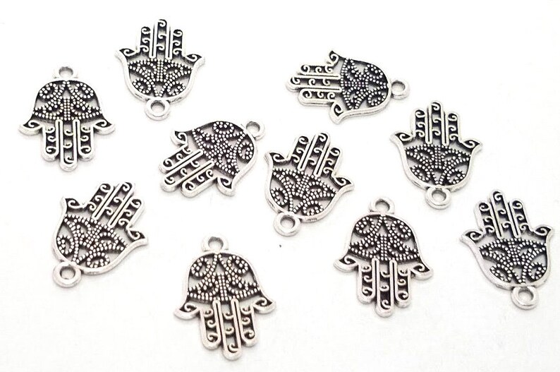 Puede incluir: Un conjunto de nueve dijes de metal plateado con forma de mano de hamsa. Cada dije presenta un dise&ntilde;o intrincado de filigrana calada con un patr&oacute;n decorativo central y una peque&ntilde;a anilla circular en la parte superior para su fijaci&oacute;n.