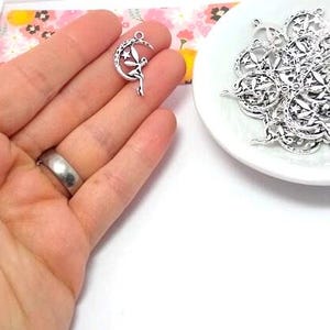 May include: A collection of silver-toned metal charms, each featuring a fairy figure seated on a crescent moon with a small star detail. The charms have a loop at the top for attachment and are presented in a small white dish with a pile of duplicates.