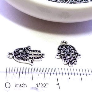 Puede incluir: Dos dijes de mano de Hamsa de metal plateado con intrincados patrones de filigrana y puntos, cada uno con una peque&ntilde;a argolla superior. Una regla de pl&aacute;stico transparente muestra que miden aproximadamente 2 cm de ancho cada uno.