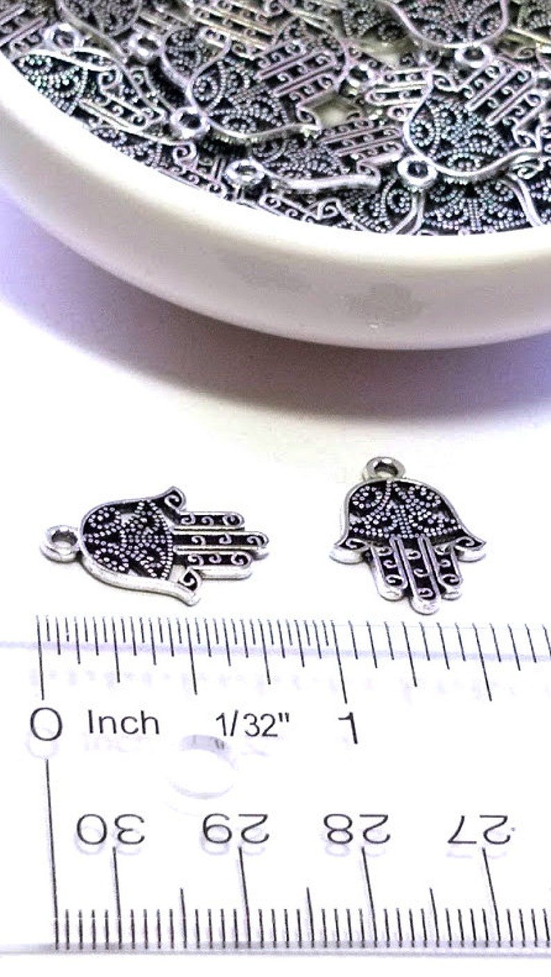 Puede incluir: Dos dijes de mano de Hamsa de metal plateado con intrincados patrones de filigrana y puntos, cada uno con una peque&ntilde;a argolla superior. Una regla de pl&aacute;stico transparente muestra que miden aproximadamente 2 cm de ancho cada uno.