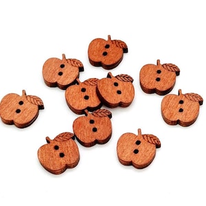 May include: A collection of ten small, brown, apple-shaped wooden buttons. Each button features a two-hole design in the center and a small leaf detail on the top right side. The buttons have a visible wood grain texture and a flat, smooth surface.