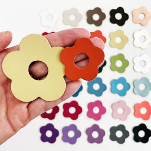 May include: A hand holds two flower-shaped leather cutouts, one yellow and one orange, with a variety of other flower cutouts in various colors scattered in the background. The cutouts have a hole in the center.