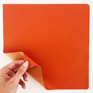 May include: A square, orange leather sheet with a folded corner revealing a tan underside. The sheet is held by a hand with manicured nails against a white background. The leather appears smooth and supple.