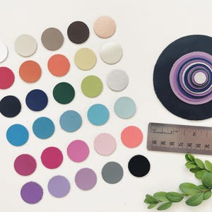 May include: A collection of colorful, circular leather swatches in various shades, including pink, blue, green, and neutral tones. A black and purple layered circle design is also present, along with a ruler showing inches. A sprig of greenery adds a natural touch.