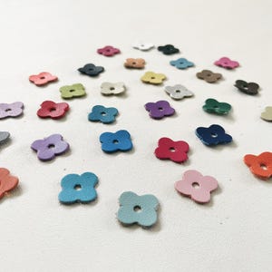 Può includere: Una collezione di piccoli ritagli di pelle a forma di fiore a quattro petali in vari colori, tra cui rosso, blu, verde e viola. I ritagli sono sparsi su una superficie bianca, creando una disposizione visivamente accattivante. Si tratta probabilmente di forniture artigianali.