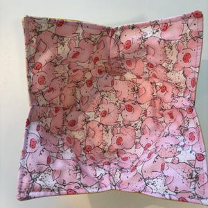 May include: A fabric bowl cozy featuring a repeating pattern of pink cartoon pigs on a white background with small black speckles. The square-shaped holder has four folded corners and visible stitching along the edges and center seams.