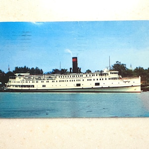 May include: A vintage postcard featuring a white steamship on blue water. The ship has a black smokestack with a red band and the text OLD BAY LINE BETWEEN NORFOLK & WASHINGTON printed on its side. The scene includes green trees in the background.