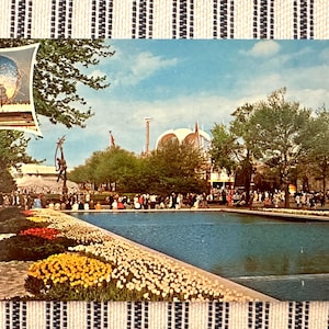 May include: Vintage postcard featuring a scenic view of the 1964-65 New York World's Fair. The image shows the Unisphere, a large pool, and crowds of people. Colorful flowers line the pool's edge, with trees and buildings in the background.