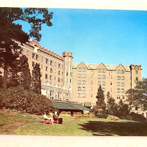 Op de afbeelding: Een vintage ansichtkaart met een groot, bruin bakstenen gebouw met een kasteelachtig ontwerp. Het gebouw heeft meerdere ramen en een groot gazon ervoor. Twee mensen zitten op rode stoelen op het gazon.