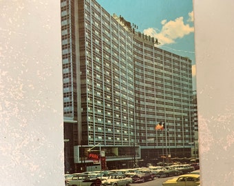 May include: Vintage postcard featuring The Statler Hilton hotel. The image shows a tall, multi-story building with numerous windows, a parking lot filled with classic cars, and a blue sky. The American flag is visible.