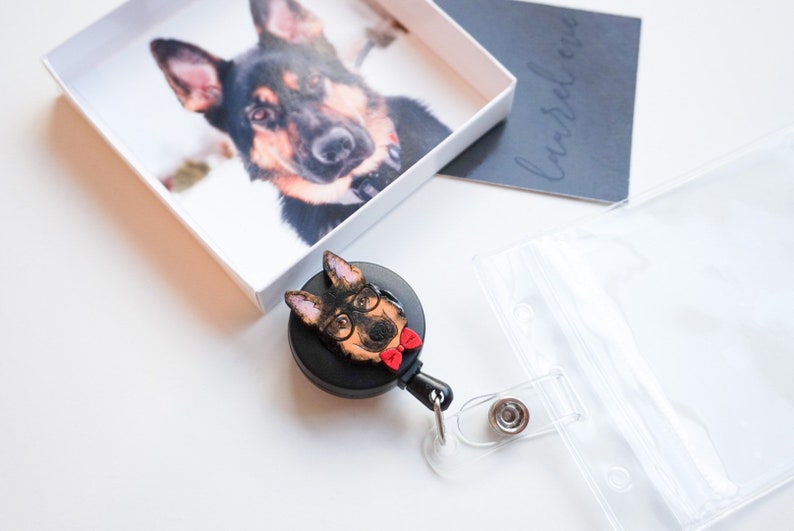 Black work badge reel with clip and clear case with a card slot. Painted wooden German Shepard dog cutout glued to badge reel. Dog has black glasses on and a red bow tie.