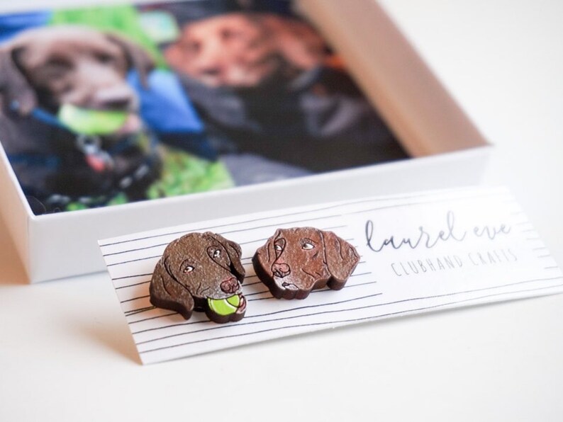 Custom pet earrings attached to a white display card sitting in front of the custom pet box lid with an image of 2 dogs inside.
One custom earring is a chocolate lab with a yellow tennis ball in mouth & the other earring is a dog with a closed-mouth.