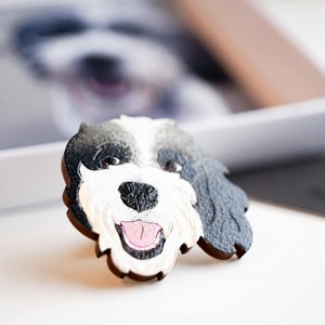 Custom pet pin of a black, white, grey dog with mouth open. Happy expression. Pin sitting in front of gift box lid with an image of the same dog inside. The pin is an identical replication of the dog image.