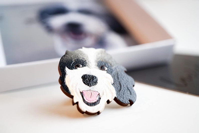 Custom pet pin of a black, white, grey dog with mouth open. Happy expression. Pin sitting in front of gift box lid with an image of the same dog inside. The pin is an identical replication of the dog image.