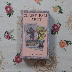 May include: A rectangular tarot card deck box with a light pink label featuring the text CLASSY PAM TAROT and Lisa Papez. The box displays two tarot cards, the Page of Cups and The Fool, against a decorative border with a floral and sun pattern.