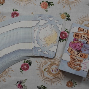 May include: A deck of Sassy Dragons Tarot cards with a light blue sky and cloud pattern, featuring a box labelled Sassy Dragons Tarot by Peggy Papez, illustrated by Andrew Rodgers and Christopher Penzal, supervised by Lion Papez with two dragons.