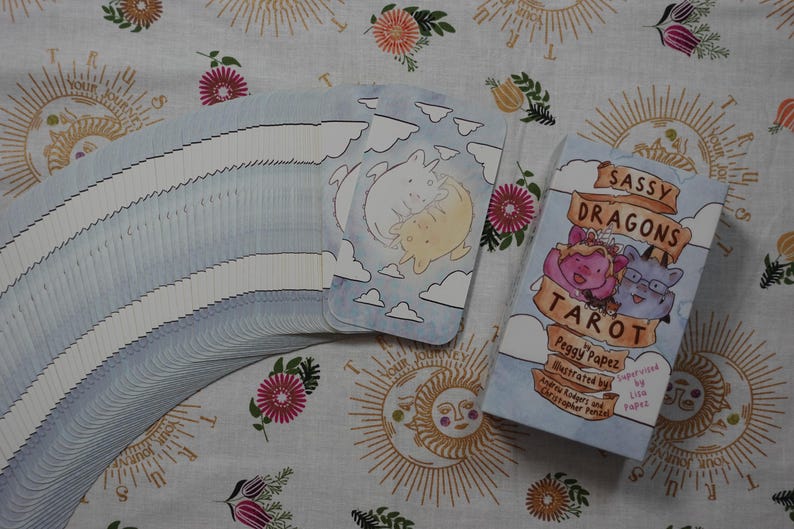 May include: A deck of Sassy Dragons Tarot cards with a light blue sky and cloud pattern, featuring a box labelled Sassy Dragons Tarot by Peggy Papez, illustrated by Andrew Rodgers and Christopher Penzal, supervised by Lion Papez with two dragons.