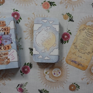 May include: A Sassy Dragons Tarot deck box, a metal card tin featuring two cartoon dragons among clouds, and a tan certificate of authenticity with the text Sassy Dragons Tarot by Peggy Papez, copy number 0159 of 1000, and a signature.