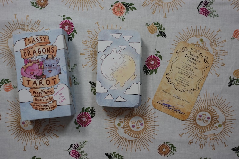 May include: A Sassy Dragons Tarot deck box, a metal card tin featuring two cartoon dragons among clouds, and a tan certificate of authenticity with the text Sassy Dragons Tarot by Peggy Papez, copy number 0159 of 1000, and a signature.