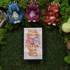May include: A Sassy Dragons Tarot deck box by Peggy Papez, illustrated by Andrew Rodgers and Christopher Papez, featuring a cartoon dragon illustration. The deck is surrounded by three small dragon figurines, a bowl of polished stones, and a plush toy.