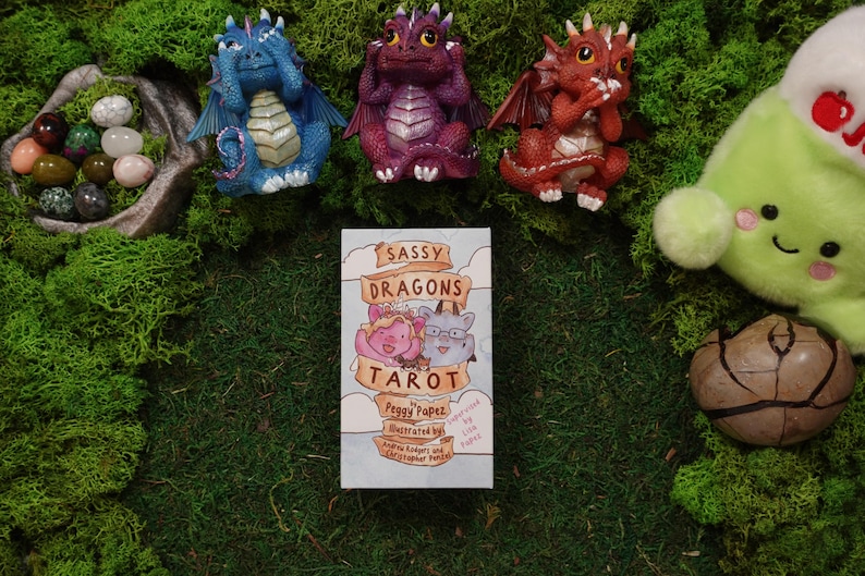 May include: A Sassy Dragons Tarot deck box by Peggy Papez, illustrated by Andrew Rodgers and Christopher Papez, featuring a cartoon dragon illustration. The deck is surrounded by three small dragon figurines, a bowl of polished stones, and a plush toy.