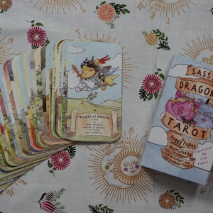 May include: A deck of Sassy Dragons Tarot cards by Peggy Papez, featuring a fan of illustrated cards and a box with the title Sassy Dragons Tarot. The top card shows a dragon knight on a horse with the text Knight of Swords. The cards have rounded corners.
