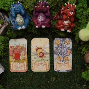 May include: Three small dragon figurines in blue, purple, and red, each covering their eyes, ears, or mouth. Below them are three tarot cards titled Six of Wands, Strength, and Wheel of Fortune, alongside a box labelled Peggy Papez Tarot.
