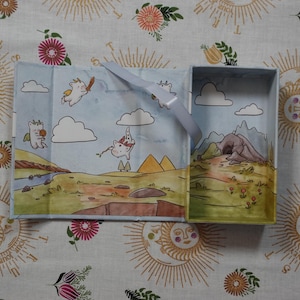 May include: A rectangular light blue box with an open lid, featuring an interior illustration of white winged creatures flying over a landscape with clouds, mountains, and a cave. A light blue ribbon is attached to the inner edge of the box.