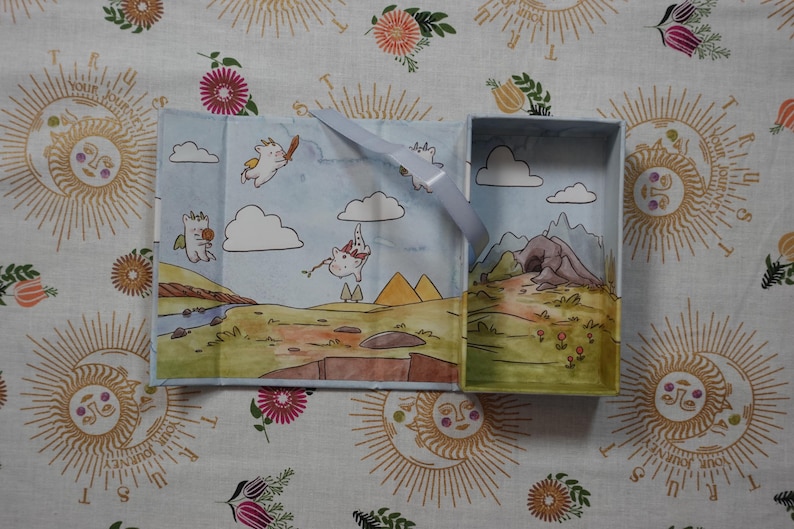 May include: A rectangular light blue box with an open lid, featuring an interior illustration of white winged creatures flying over a landscape with clouds, mountains, and a cave. A light blue ribbon is attached to the inner edge of the box.