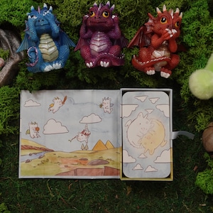 May include: Three small dragon figurines in blue, purple, and red, each covering their eyes, ears, or mouth. Below them is a rectangular box featuring a cartoon illustration of two white creatures and a landscape with clouds and pyramids.
