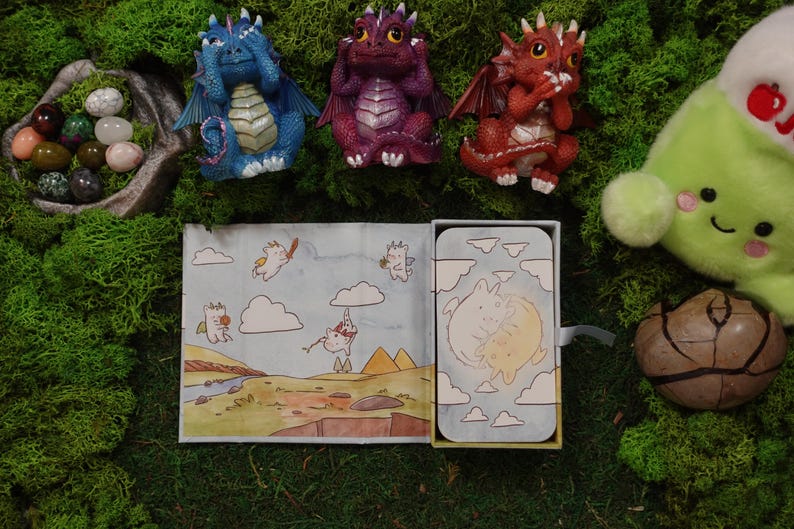 May include: Three small dragon figurines in blue, purple, and red, each covering their eyes, ears, or mouth. Below them is a rectangular box featuring a cartoon illustration of two white creatures and a landscape with clouds and pyramids.