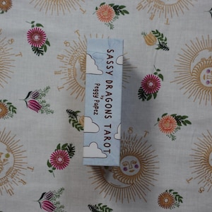 May include: A rectangular tarot deck box with a light blue background and white cloud illustrations. The text on the side reads Sassy Dragons Tarot by Peggy Papez. The box is positioned vertically against a white fabric with sun and floral patterns.