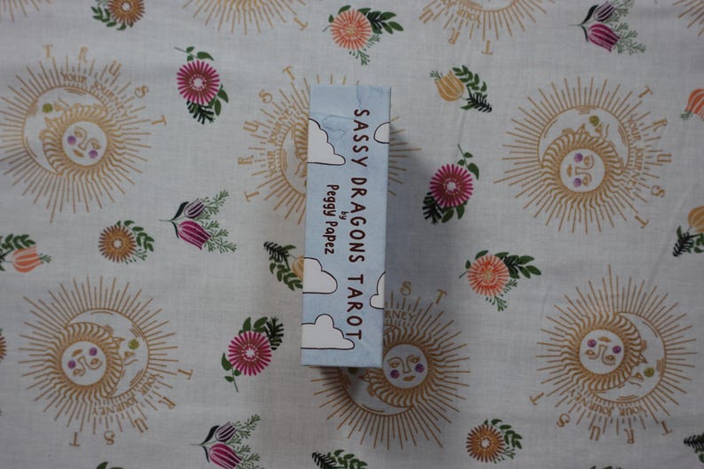 May include: A rectangular tarot deck box with a light blue background and white cloud illustrations. The text on the side reads Sassy Dragons Tarot by Peggy Papez. The box is positioned vertically against a white fabric with sun and floral patterns.