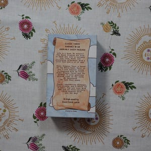 May include: A rectangular tarot card deck box with a light blue background and white clouds. The front features a tan scroll graphic with black text that reads Classic Tarot Guidance in an Adorably Sassy Package, detailing the deck's contents.