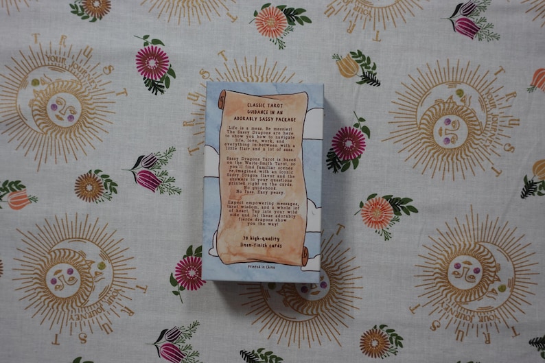 May include: A rectangular tarot card deck box with a light blue background and white clouds. The front features a tan scroll graphic with black text that reads Classic Tarot Guidance in an Adorably Sassy Package, detailing the deck's contents.