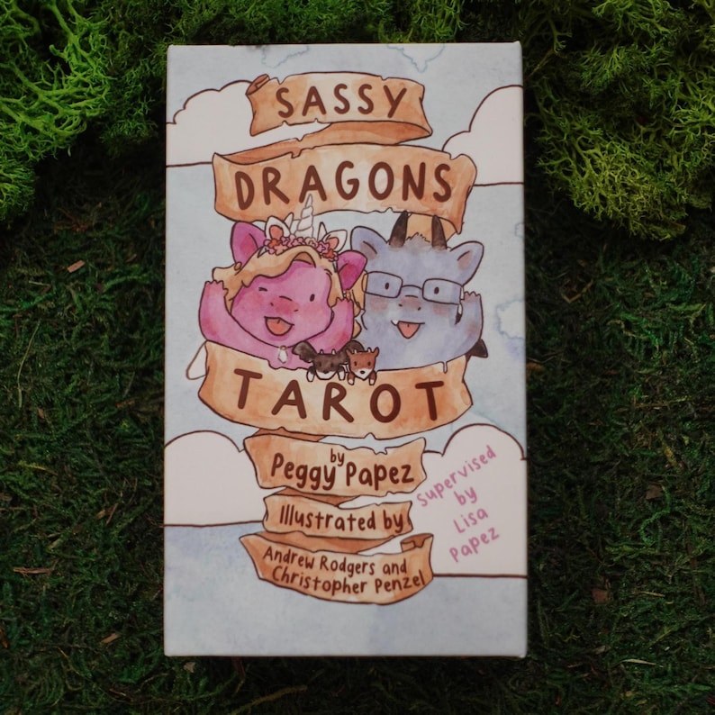 May include: A rectangular tarot deck box featuring the title Sassy Dragons Tarot on a tan ribbon banner. The illustration shows a pink dragon with a unicorn horn and a grey dragon wearing glasses, with text credits for Peggy Papez, Lisa Papez, Andrew Rodgers, and Christopher Penzel.