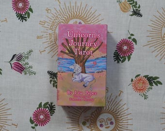 May include: A rectangular tarot card deck box titled Unicorn's Journey Tarot by Lisa Papez and illustrated by Francesca Paiocchi. The box features a pink and orange sunset sky, a brown tree, and a white unicorn resting on a sandy beach by blue water.