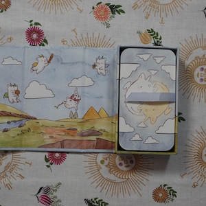 May include: A rectangular box set featuring a deck of cards with a light blue sky and cloud illustration. The open lid displays a landscape with cartoon creatures, while the card deck is secured with a light blue paper band showing two figures.