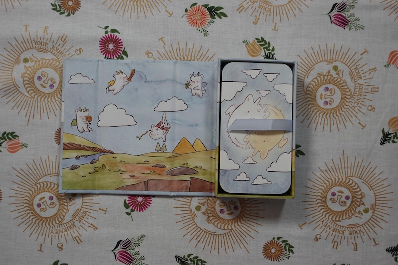 May include: A rectangular box set featuring a deck of cards with a light blue sky and cloud illustration. The open lid displays a landscape with cartoon creatures, while the card deck is secured with a light blue paper band showing two figures.