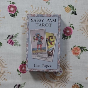 May include: A rectangular tarot card deck box with a light blue cover featuring the text SASSY PAM TAROT and Lisa Papez. The box displays two tarot cards on the front, the Page of Cups and the Fool, against a floral patterned border.