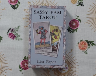 Könnte beinhalten: Eine rechteckige Tarotkartenschachtel mit hellblauem Deckel, auf dem der Text SASSY PAM TAROT und Lisa Papez steht. Die Schachtel zeigt auf der Vorderseite zwei Tarotkarten, den Buben der Kelche und den Narren, vor einem floralen Rand.