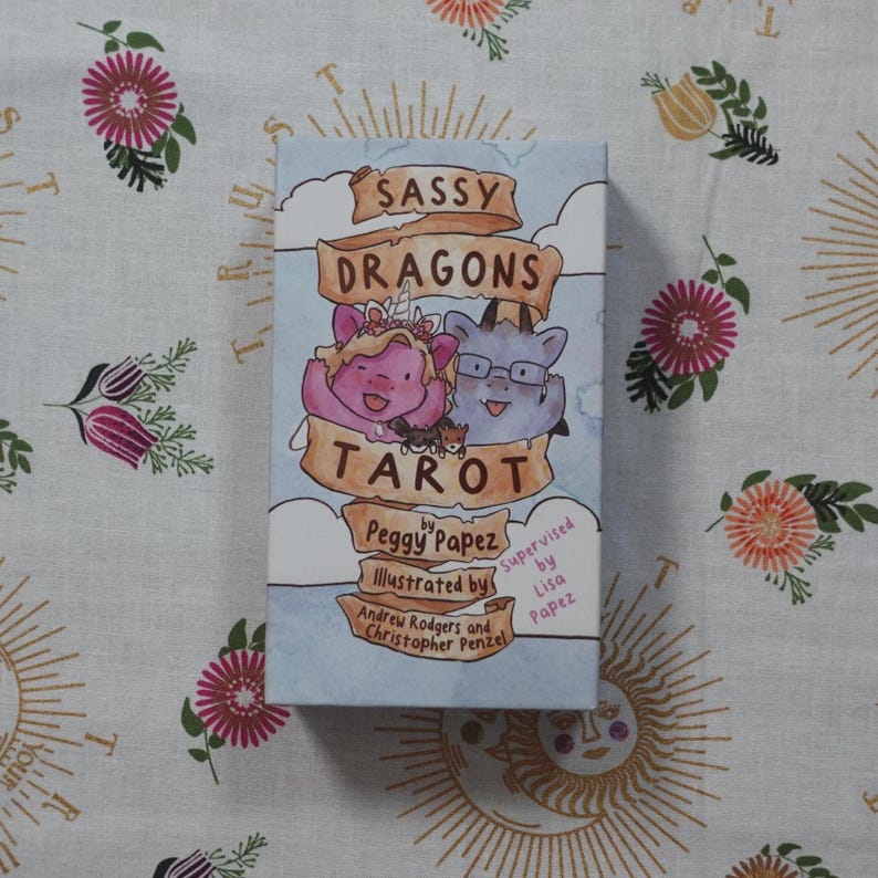 May include: A rectangular tarot deck box titled Sassy Dragons Tarot by Peggy Papez. The cover features a light blue sky background with white clouds and a central illustration of two cartoon dragons, one pink and one blue, framed by tan banners.