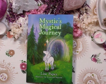 May include: A book titled Mystic's Magical Journey by Lisa Papez, illustrated by Francesca Paiocchi. The cover features a white unicorn standing in a green forest with tall pine trees and a glowing portal, with a small orange butterfly near its horn.
