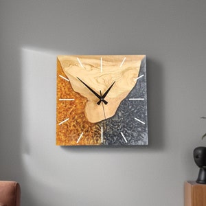 Puede incluir: Reloj de pared cuadrado con un diseño único. La esfera presenta un patrón de vetas de madera natural, complementado con secciones de resina dorada y gris oscuro. Líneas blancas marcan las horas, y las manecillas negras indican la hora.